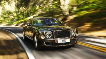 Bentley Mulsanne front view, featuring Matt Black matrix grille and Bentley Radiator Mascot, driving along a road with trees in backdrop. 