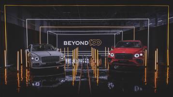 Bentley Bentayga in St James red and Bentley Flying Spur Speed Edition 12 in Tyrolean, front side angled view parked indoors with Beyond 100 neon sign in background