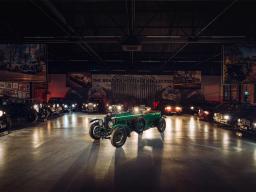 Top side angled view of Bentley blower parked in museum with other Bentley's legacy model around in back ground with headlamps switched on.
