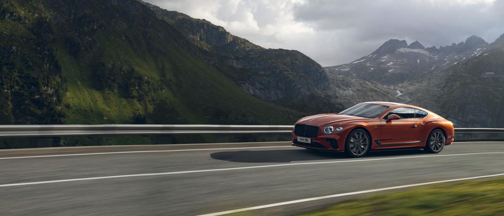 Side view of Bentley Continental GT Speed W12 in Orange Flame colour driving along a highway with mountains in backdrop. 