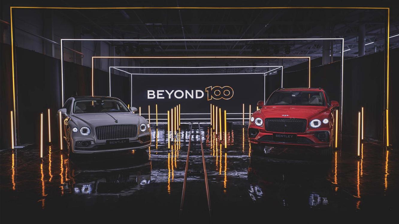 Bentley Bentayga in St James red and Bentley Flying Spur in Titan Grey, front side angled view parked indoors with Beyond 100 neon sign in background