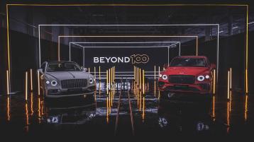 Bentley Bentayga in St James red and Bentley Flying Spur in Titan Grey, front side angled view parked indoors with Beyond 100 neon sign in background