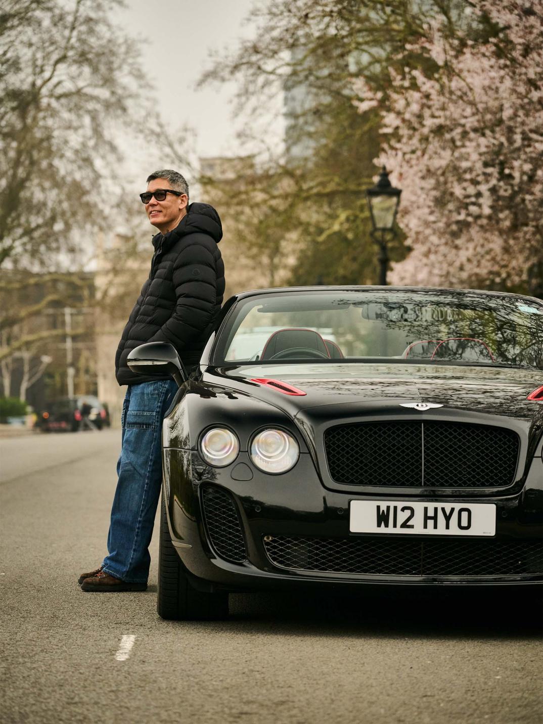Chart Chirathivat leaning against his Bentley Supersports ISR, which is parked at the side of a road.