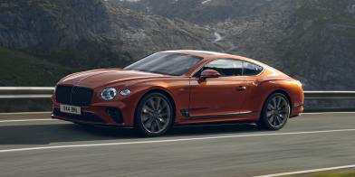Side angled view of Bentley Continental GT Speed in Orange Flame, driving along highway with mountains in backdrop. 