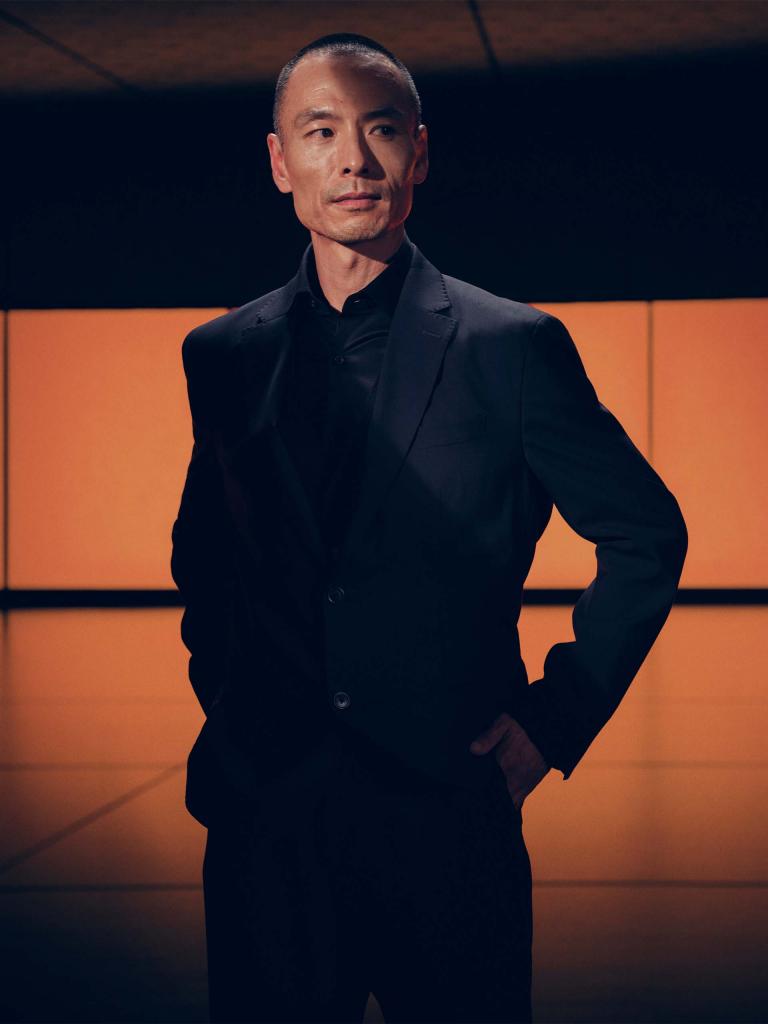 A person/model/man/ in black sports jacket, with hands in pocket posing for a photograph.