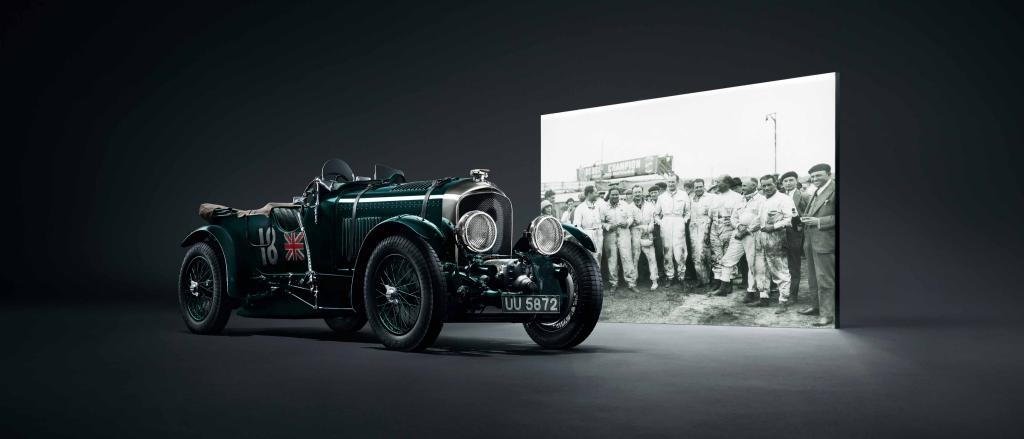 Side angled view of Bentley Blower Jnr in Verdant Green view, parked infront of projected black and white photograph of Bentley Boys