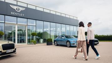 Two individuals heading into CW1 House. Home of Bentley, featuring panoramic glass view and Bentley Flying Spurs in view.