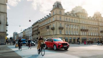 Side angled view of Bentley Bentayga EWB in St James Red colour driving along an Urban setting with bicyclists in view.