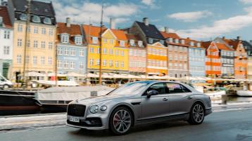 Side view of Bentley Flying Spur S in Cambrian Grey driving on road, with colourful Scandinavian architecture in view.