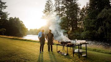 Two individuals standing infront of grille in an outdoor setting, with river running in background.