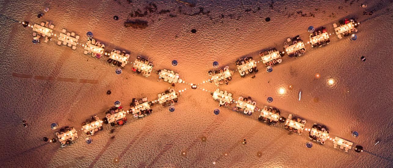 Birds Eye view of People dining with tables set in X shape in sandy terrain.