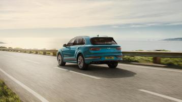 Rear view of Bentley Bentayga Azure in Topaz Blue colour with black painted shark fin aerial and fixed rear spoiler and jewel cut taillamps driving along sea side.