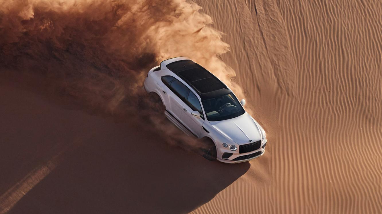 Aerial view of Bentayga Speed in Snow Quartz by Mulliner exterior paint with the Bentayga Styling Specification and 23" Directional Wheels driving down a sand dune