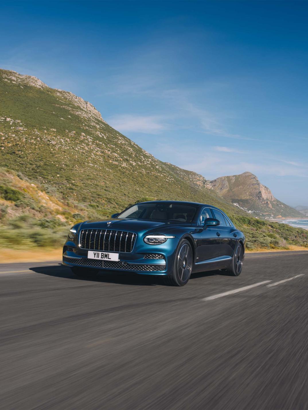 Front three-quarter shot of the Bentley Flying Spur Azure in Dark Teal with Bright Chrome Exterior detailing on a sunny road.