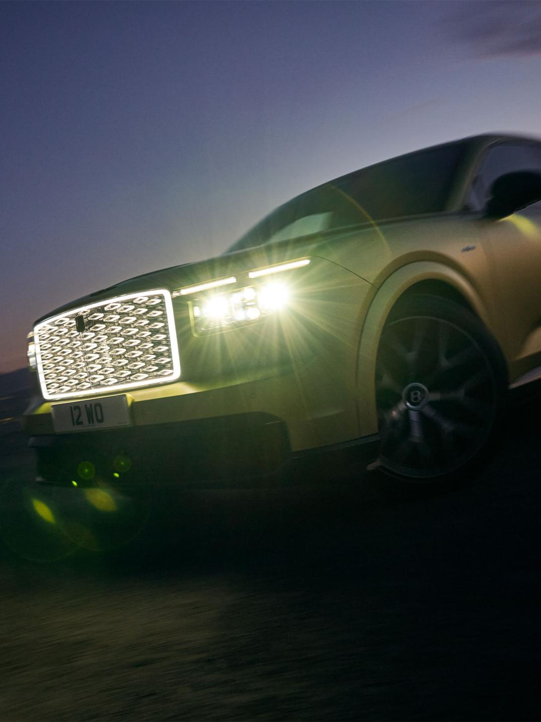Front close-up driving shot of the Bentley Torcal S with the headlights illuminated.