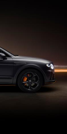 Light grey satin Bentley Bentayga S Black Edition, driving on a bridge in an urban environment featuring 22 inch S directional wheel - Black Painted and Polished as well as Mandarin accents on the styling kit and break callipers