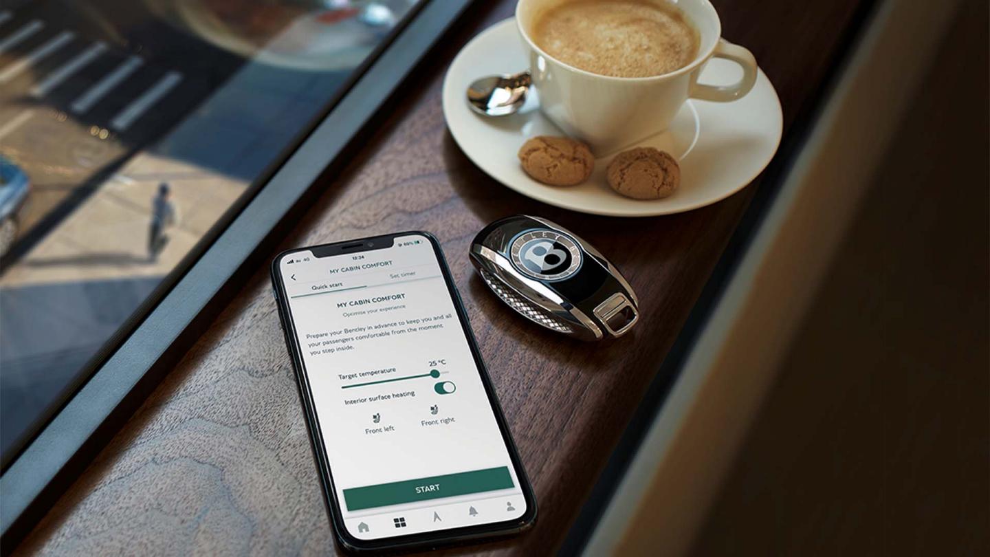 Phone with the My Bentley app open, next to a Bentley key and a cup of coffee, overlooking a street.