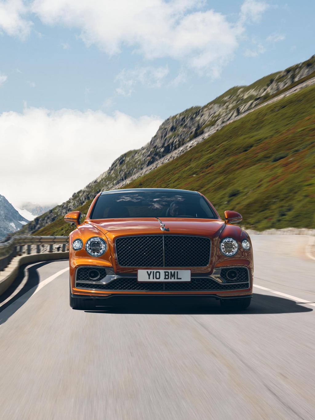 Front view of Bentley Flying Spur Speed W12, in Orange flame driving along hills, featuring chrome matrix grille and radiator mascot – black polished 