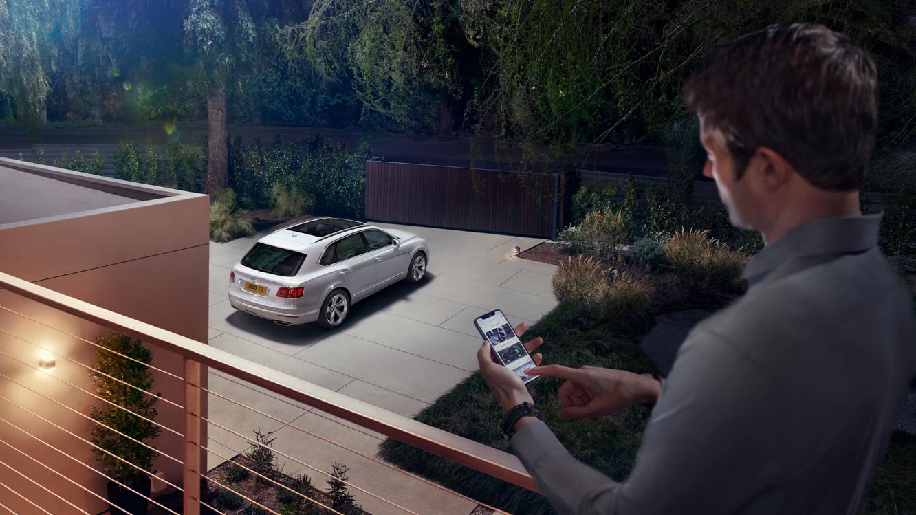 A person using My Bentley App from house terrace connected to Bentley Bentayga parked in drive.