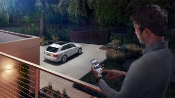 A person using My Bentley App from house terrace connected to Bentley Bentayga parked in drive.