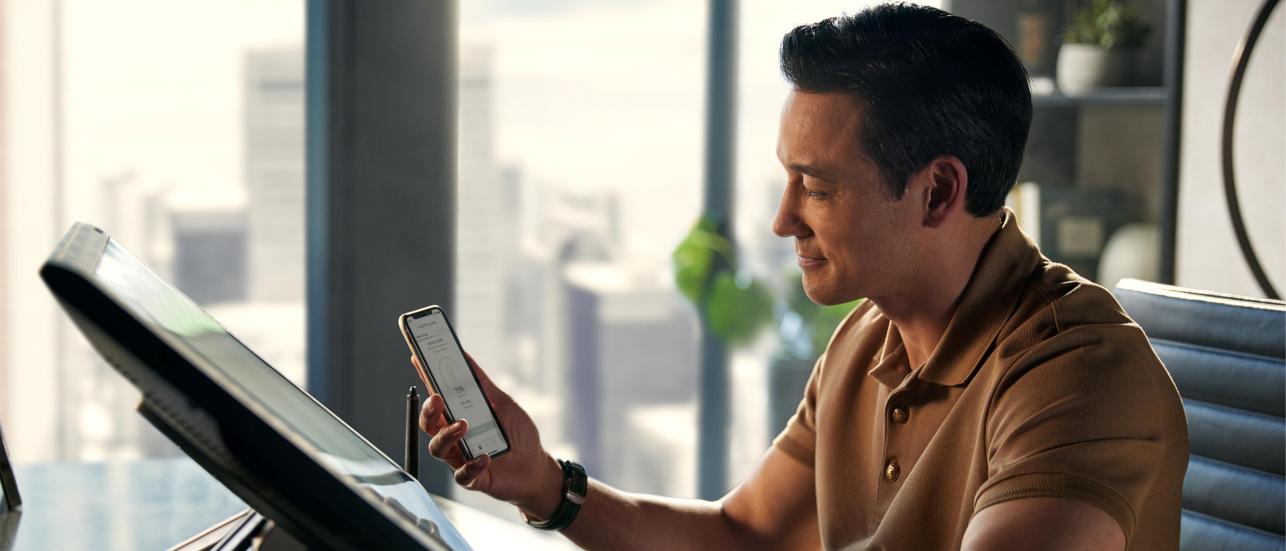 A person infront of deskop screen looking at phone displaying My Bentley App.